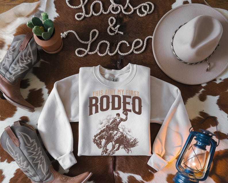 Peut inclure: Un sweat-shirt blanc avec un graphique brun d'un cowboy chevauchant un bronco. Le texte "This ain't my first rodeo" est imprim&eacute; en brun au-dessus du graphique.