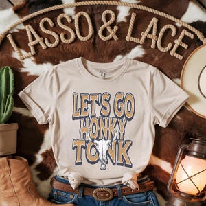 May include: A beige t-shirt with the text "Let's Go Honky Tonk" in yellow and blue lettering. A stylized skull with horns is in the center of the text.