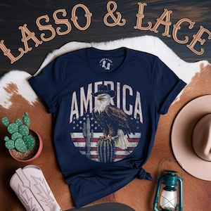 May include: Navy blue t-shirt with a distressed graphic of an eagle wearing a cowboy hat and perched on a cactus. The eagle is in front of an American flag with the word "AMERICA" in white text above it.