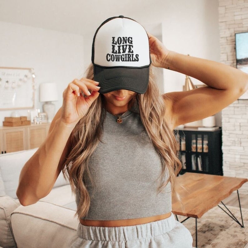 Op de afbeelding: Een witte en zwarte truckercap met de tekst "Long Live Cowgirls" in zwarte borduurwerk. De pet wordt gedragen door een persoon met lang blond haar.