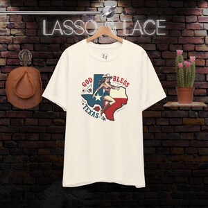K&ouml;nnte beinhalten: Ein wei&szlig;es T-Shirt mit einer Grafik im Vintage-Stil einer Frau im Cowboyhut, die auf einer texanischen Umrissform steht. Die Grafik lautet "God Bless Texas" in Rot, Wei&szlig; und Blau.