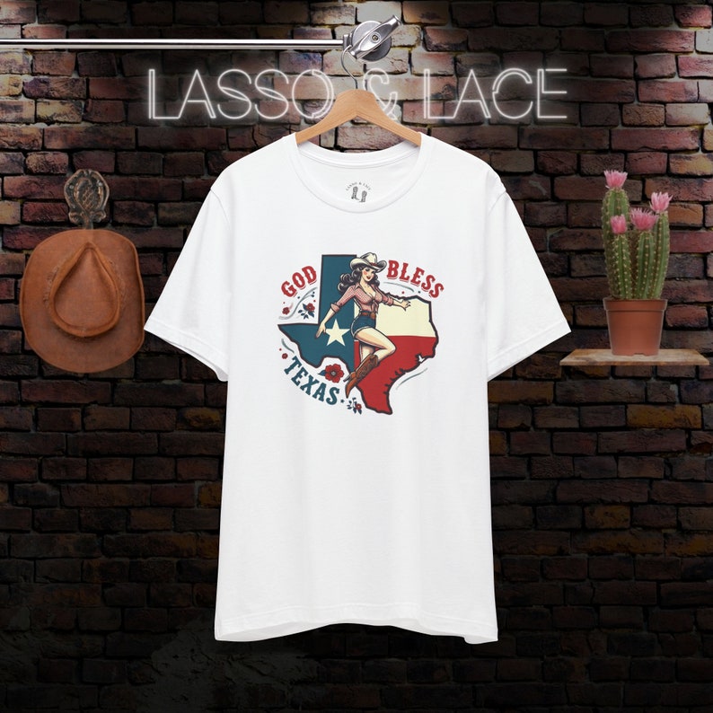 K&ouml;nnte beinhalten: Wei&szlig;es T-Shirt mit einem Grafikdruck einer Frau in einem Cowboyhut, die auf einer Umrisskarte von Texas steht, mit den Worten "God Bless Texas" darauf gedruckt.