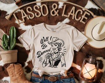 Op de afbeelding: Een beige T-shirt met een zwart-witte illustratie van een cowgirl die een glas vasthoudt. De tekst "Well! Be Damned" staat op het shirt gedrukt.