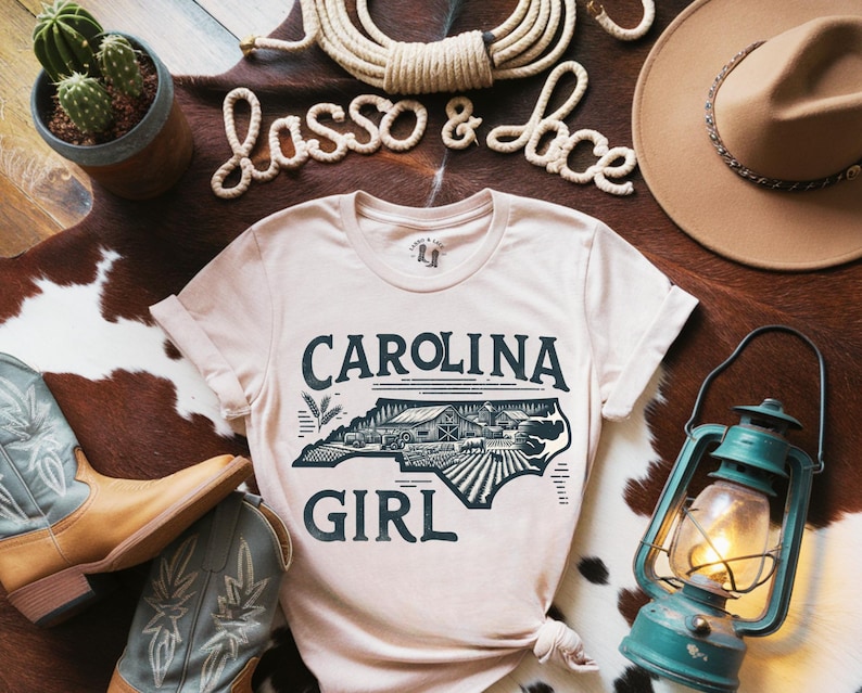May include: A light beige t-shirt with a graphic of the state of North Carolina and the text "Carolina Girl". The graphic is in black and white and features a farmhouse and fields.