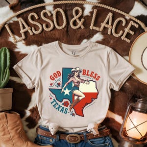 K&ouml;nnte beinhalten: Ein beiges T-Shirt mit einer Vintage-Illustration einer Frau in einem Cowboyhut und Stiefeln, die auf einer texanischen Umrissform steht. Der Text "God Bless Texas" ist &uuml;ber der Illustration gedruckt.
