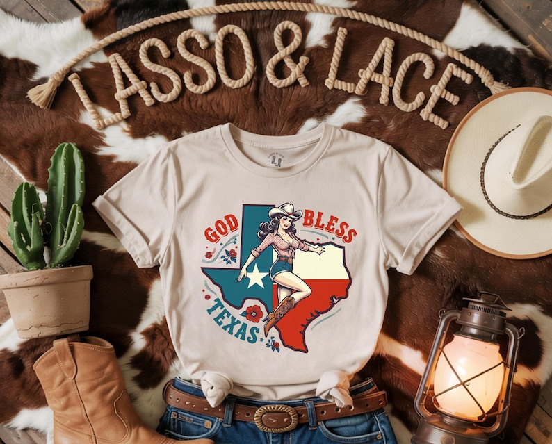 K&ouml;nnte beinhalten: Ein beiges T-Shirt mit einer Vintage-Illustration einer Frau in einem Cowboyhut und Stiefeln, die auf einer texanischen Umrissform steht. Der Text "God Bless Texas" ist &uuml;ber der Illustration gedruckt.