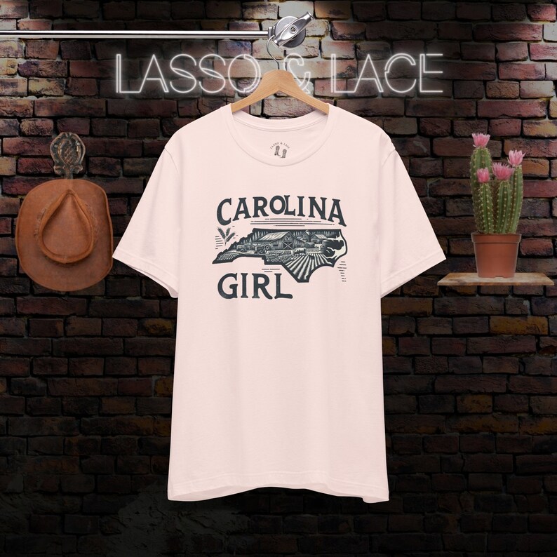 May include: A pink t-shirt with a black and white graphic of the state of North Carolina. The graphic includes the text "CAROLINA GIRL".