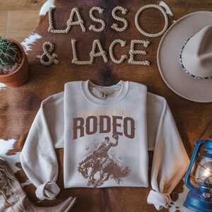 Peut inclure: Un sweat-shirt beige avec le texte "This ain't my first rodeo" en lettres brunes. Le sweat-shirt pr&eacute;sente un graphique d'un cow-boy chevauchant un bronco qui se cabre.