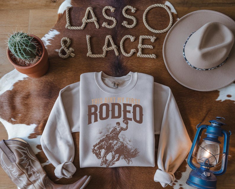 Peut inclure: Un sweat-shirt beige avec le texte "This ain't my first rodeo" en lettres brunes. Le sweat-shirt pr&eacute;sente un graphique d'un cow-boy chevauchant un bronco qui se cabre.