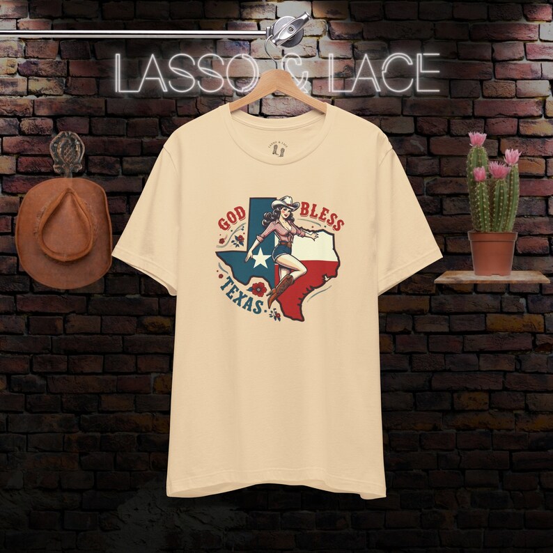 K&ouml;nnte beinhalten: Ein beiges T-Shirt mit einem Grafikdruck einer Frau in einem Cowboyhut, die auf einer stilisierten Umrandung des Bundesstaates Texas steht. Die Grafik enth&auml;lt den Text "God Bless Texas" und ist in den Farben Rot, Wei&szlig; und Blau gehalten.