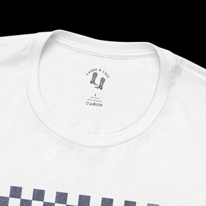 May include: White t-shirt with a black and white checkered pattern on the bottom. The neck label reads "LASSO & LACE" with a graphic of two cowboy boots. The label also includes "100% Cotton Made in Bangladesh" and washing instructions.