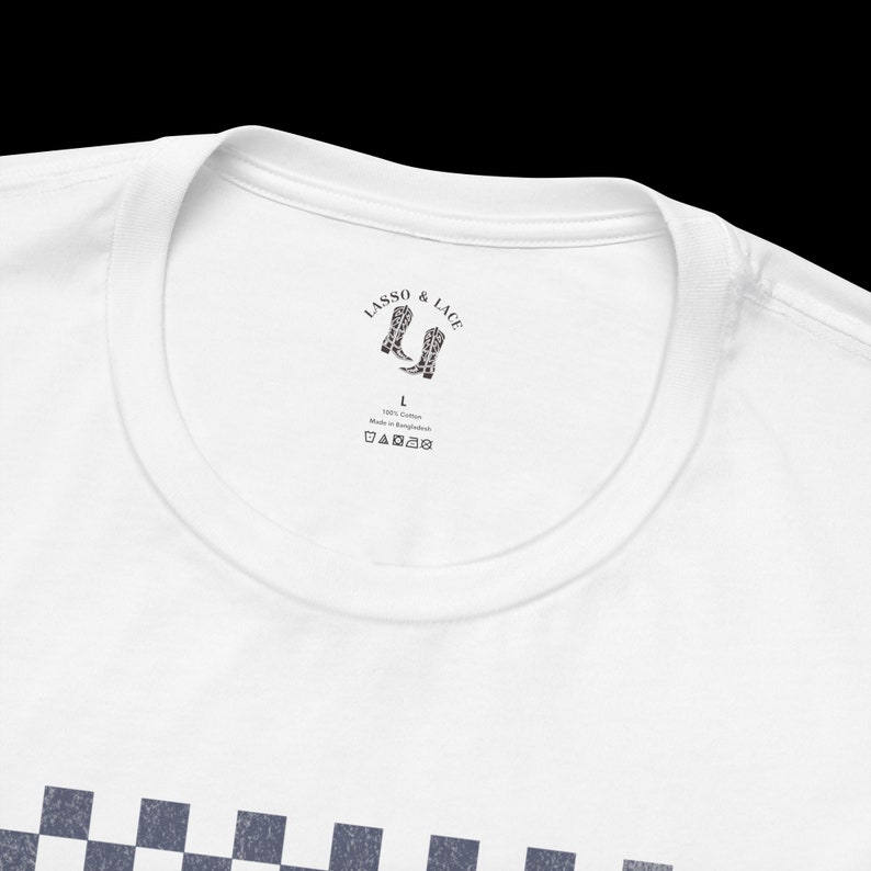 May include: White t-shirt with a black and white checkered pattern on the bottom. The neck label reads "LASSO & LACE" with a graphic of two cowboy boots. The label also includes "100% Cotton Made in Bangladesh" and washing instructions.