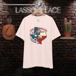 K&ouml;nnte beinhalten: Rosafarbenes T-Shirt mit einem Grafikdruck einer Frau in einem Cowboyhut, die auf einer texanischen Umrissform steht, mit den Worten "God Bless Texas" drumherum.