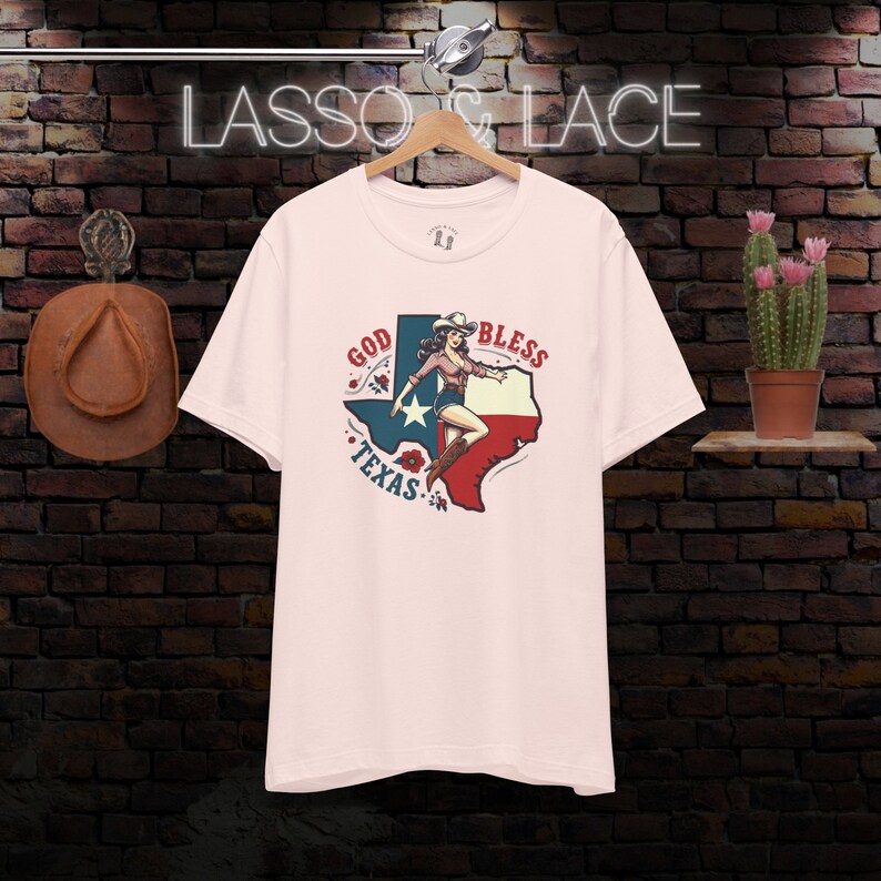 K&ouml;nnte beinhalten: Rosafarbenes T-Shirt mit einem Grafikdruck einer Frau in einem Cowboyhut, die auf einer texanischen Umrissform steht, mit den Worten "God Bless Texas" drumherum.