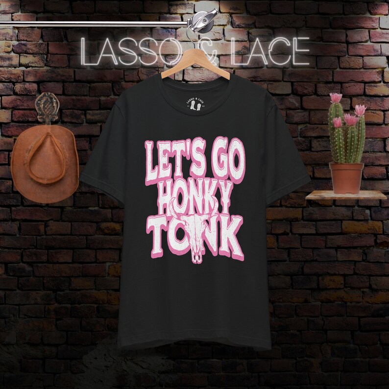 May include: Black t-shirt with a pink graphic that says "Let's Go Honky Tonk" with a pink stylized bull skull.