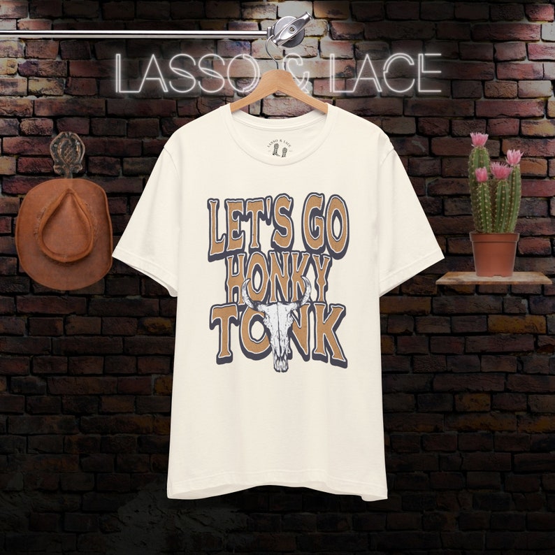 May include: A white t-shirt with a brown graphic that says "Let's Go Honky Tonk" with a stylized bull skull in the middle of the text.