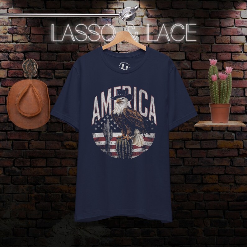 May include: A navy blue t-shirt with a vintage-style graphic of an eagle wearing a cowboy hat, perched on a cactus, with the word "AMERICA" in a circle around it. The eagle and cactus are in front of a faded American flag.