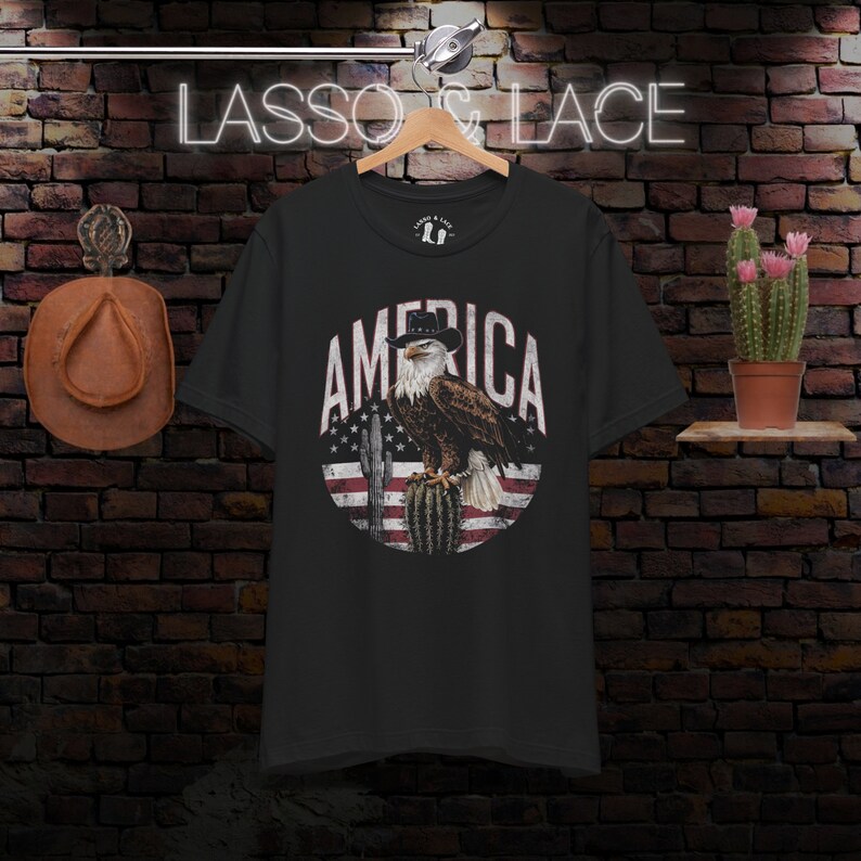 May include: Black t-shirt with a vintage-style graphic of an eagle wearing a cowboy hat, perched on a cactus with the American flag in the background. The text "AMERICA" is written in a circular design around the eagle.