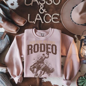 Peut inclure: Sweat-shirt rose avec un graphique marron d'un cowboy &agrave; cheval. Le graphique dit "This is my first rodeo".