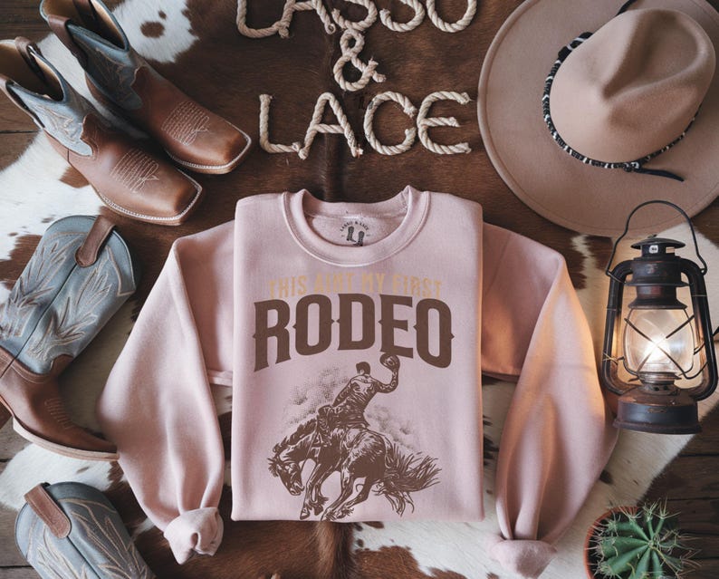 Peut inclure: Sweat-shirt rose avec un graphique marron d'un cowboy &agrave; cheval. Le graphique dit "This is my first rodeo".