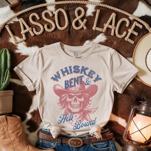 May include: A beige t-shirt featuring a vintage-style graphic of a skull wearing a cowboy hat. The graphic reads "Whiskey Bent & Hell Bound".