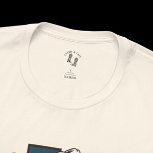 K&ouml;nnte beinhalten: Wei&szlig;es T-Shirt mit einem schwarz-wei&szlig;en Logo, das "Lasso & Lace" mit einem Paar Cowboystiefeln sagt. Das Logo befindet sich auf einem wei&szlig;en Etikett, das in den Nacken des Shirts eingen&auml;ht ist. Das Shirt ist Gr&ouml;&szlig;e L und aus 100% Baumwolle.