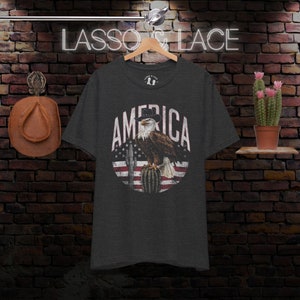 May include: A dark heather grey t-shirt with a vintage-style graphic of an eagle wearing a cowboy hat perched on a cactus. The eagle is in front of a red, white, and blue American flag. The text "AMERICA" is written in a white, distressed font around the eagle.