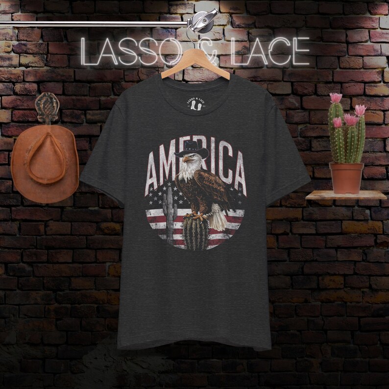 May include: A dark heather grey t-shirt with a vintage-style graphic of an eagle wearing a cowboy hat perched on a cactus. The eagle is in front of a red, white, and blue American flag. The text "AMERICA" is written in a white, distressed font around the eagle.