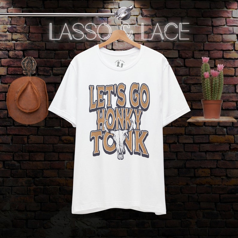 May include: White t-shirt with a brown graphic that says "Let's Go Honky Tonk" with a stylized bull skull in the middle of the text.