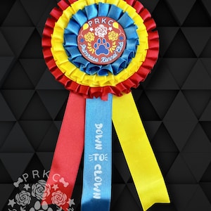 Könnte beinhalten: Eine rote, gelbe und blaue Rosette mit einem weißen Band, auf dem "Down to Clown" steht. Die Rosette hat einen Mittelkreis mit dem Text "PRKC Primrose Kennel Club" und einen Pfotenabdruck.