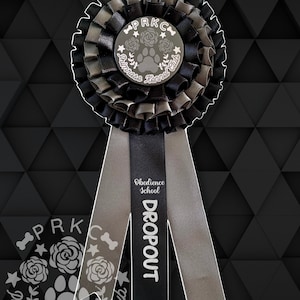 Op de afbeelding: Zwarte en grijze rozet met een zilveren pootprint en de tekst "PRKC Primrose Kennel Club" en een zwart lint met de tekst "Obedience School DROPOUT"