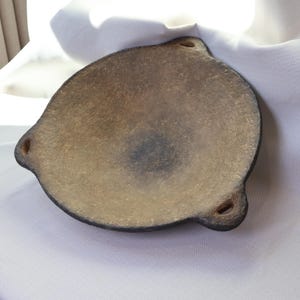 May include: A handmade, ceramic serving dish with a unique, organic shape. The dish has a light brown interior and a dark brown rim. It features three small, rounded handles and a slightly textured surface.