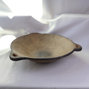 May include: A handmade, oval-shaped ceramic bowl with a textured, light brown interior and a dark rim. The bowl has two small, rounded handles and sits on a white surface. The bowl is likely used for serving food.
