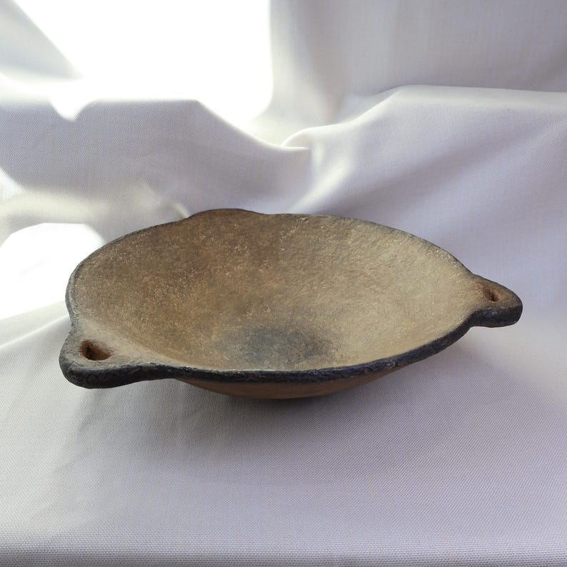 May include: A handmade, oval-shaped ceramic bowl with a textured, light brown interior and a dark rim. The bowl has two small, rounded handles and sits on a white surface. The bowl is likely used for serving food.
