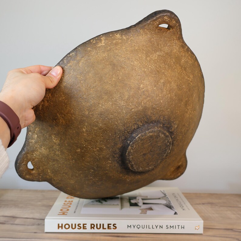 May include: A large, antique-style, brown and gold bowl with a textured surface. The bowl has two small holes on the top and a circular base. The bowl is resting on a book titled "HOUSE RULES" by Myquillyn Smith.