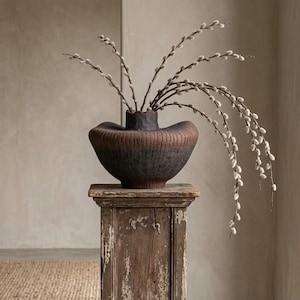 May include: A dark brown and black textured ceramic vase with a wide, flared rim and vertical ribbed detailing. The vase contains several long, slender branches with small, fuzzy white pussy willow buds that arch gracefully outward.