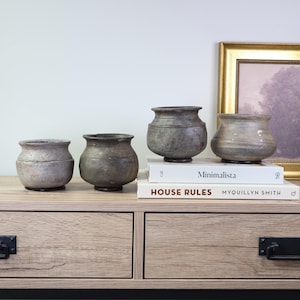 May include: Four antique, weathered ceramic pots in varying sizes and shades of gray and brown are displayed on a light wood surface. The pots have rounded bodies and slightly flared rims. Books with the titles "Minimalista" and "HOUSE RULES" are visible.