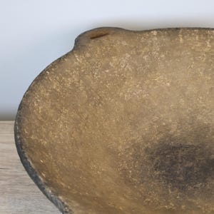 May include: A handcrafted, textured bowl with a rustic aesthetic. The bowl features a rounded shape with a slightly irregular rim and a small handle-like detail. The color is a blend of tan and brown, with darker shading.