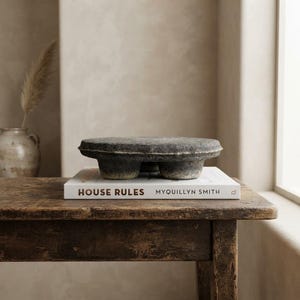 May include: A circular, dark gray, textured stone pedestal with three short, thick legs sits atop a white hardcover book titled House Rules by Myquillyn Smith. The object has a rough, weathered surface and a flat top, resting on a dark wooden table.