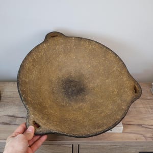 May include: A large, round, textured bowl with a light brown interior and a dark brown rim. The bowl has three small, rectangular cutouts on the rim. The bowl is sitting on a wooden surface.