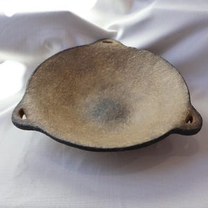 May include: A handmade, shallow, ceramic bowl with a textured, speckled surface. The bowl is primarily tan with darker brown edges and small loop handles on either side. The bowl has a unique, organic shape.