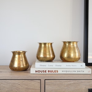 May include: Three antique brass vases of varying sizes sit atop a stack of books on a wooden cabinet. The vases have a rounded shape with a flared rim and horizontal ridges. The books read "House Love Home" and "House Rules".