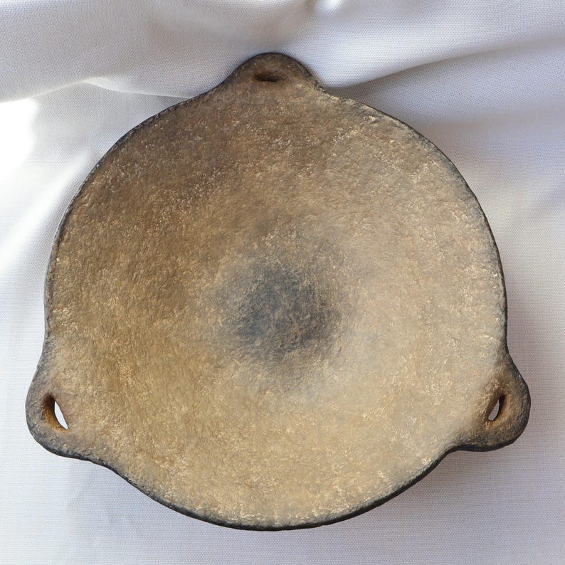 May include: A handmade, round, shallow dish with a textured, light brown surface and a dark brown rim. The dish has three rounded extensions, each with a small hole. The center of the dish is a darker shade of brown.