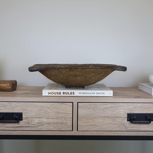 May include: A decorative, textured, brown bowl with a dark rim sits atop a book titled "HOUSE RULES" on a wooden table. The bowl has a unique, organic shape and is a stylish home decor item.