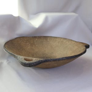 May include: A handcrafted, oval-shaped bowl with a textured, light brown interior and a dark gray rim. The bowl features small, rounded handles on each side. The overall design suggests a rustic or artisanal aesthetic.