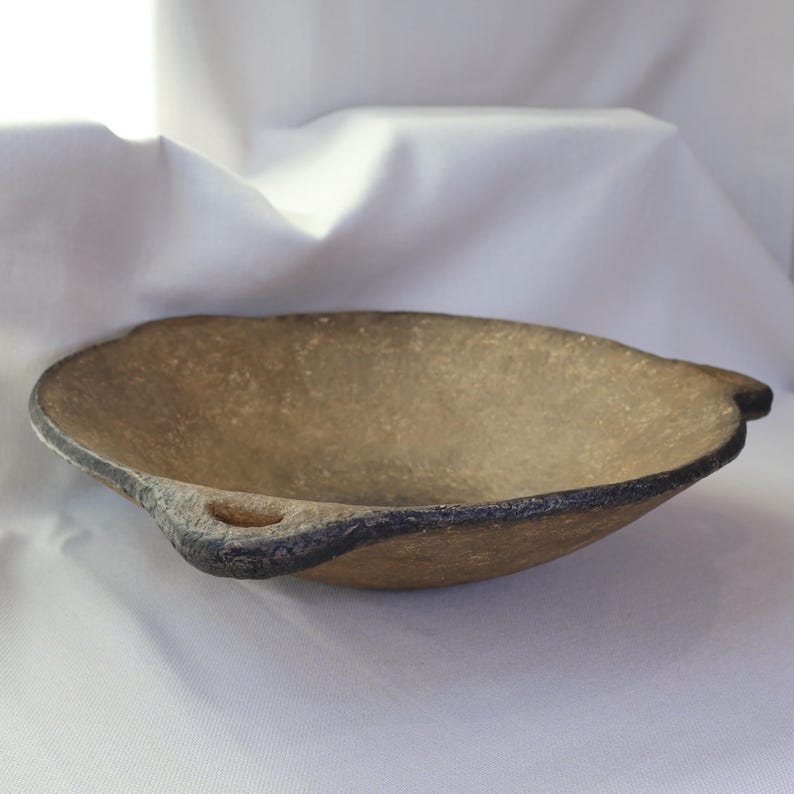 May include: A handcrafted, oval-shaped bowl with a textured, light brown interior and a dark gray rim. The bowl features small, rounded handles on each side. The overall design suggests a rustic or artisanal aesthetic.