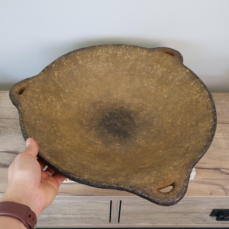 May include: A large, oval-shaped, brown bowl with a textured surface. The bowl has three small, rounded handles and a dark, circular area in the center. The bowl is made of a material that appears to be stone or ceramic.