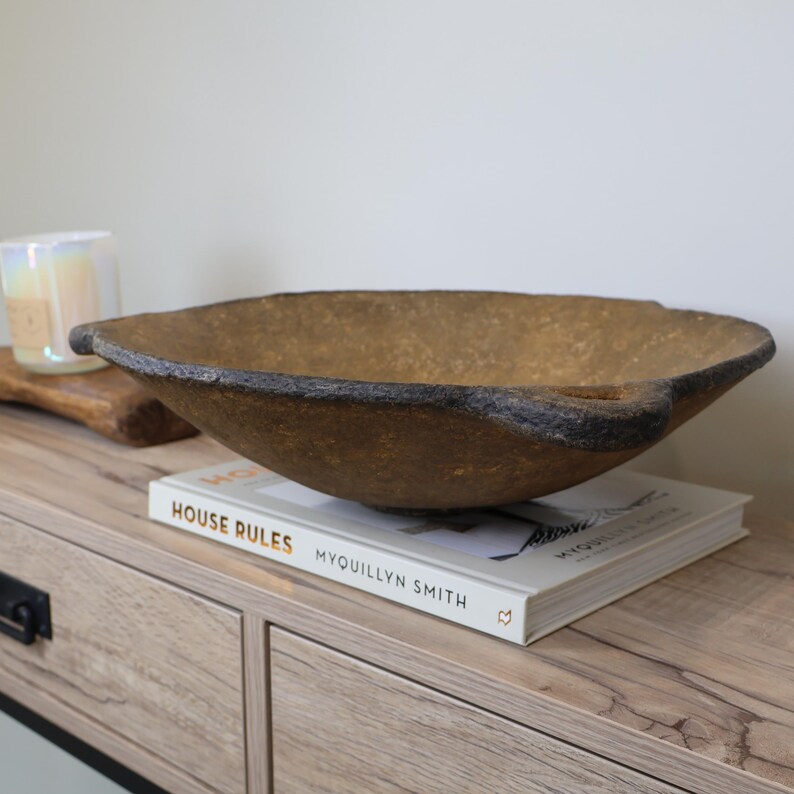 May include: A large, textured, brown decorative bowl with dark edges and two handles. The bowl sits on a white book with the title "HOUSE RULES" and the author's name "MYQUILLYN SMITH". The bowl is placed on a light-colored wooden surface.