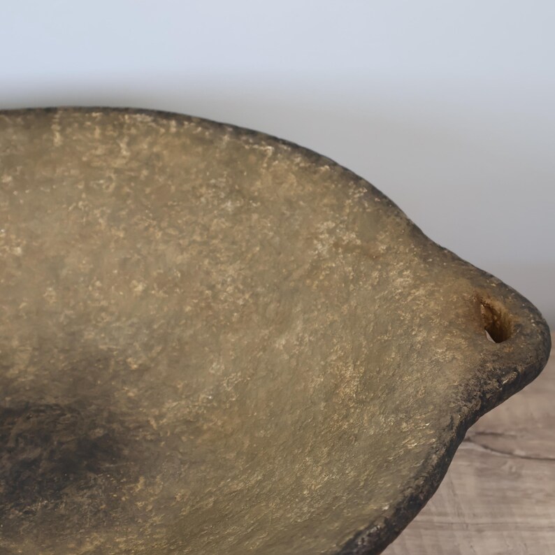 May include: A rustic, oval-shaped serving bowl with a textured, mottled surface in shades of tan and brown. The bowl has a slightly raised rim and a small hole on one side, suggesting a handle. The overall aesthetic is vintage or antique.