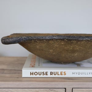 May include: A textured, gold-colored bowl with a dark rim and a handle. The bowl is resting on a book titled "HOUSE RULES" with the author's name, Myquillyn, visible. The bowl is a decorative piece.